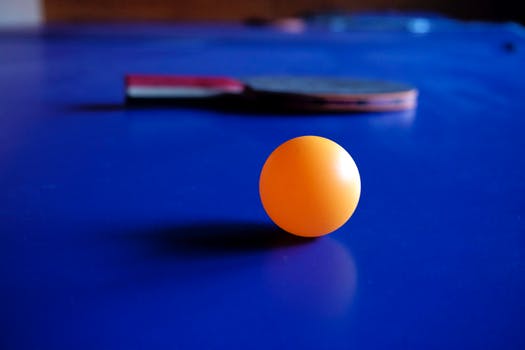 Bright orange ping pong ball on a bright blue table with a paddle.