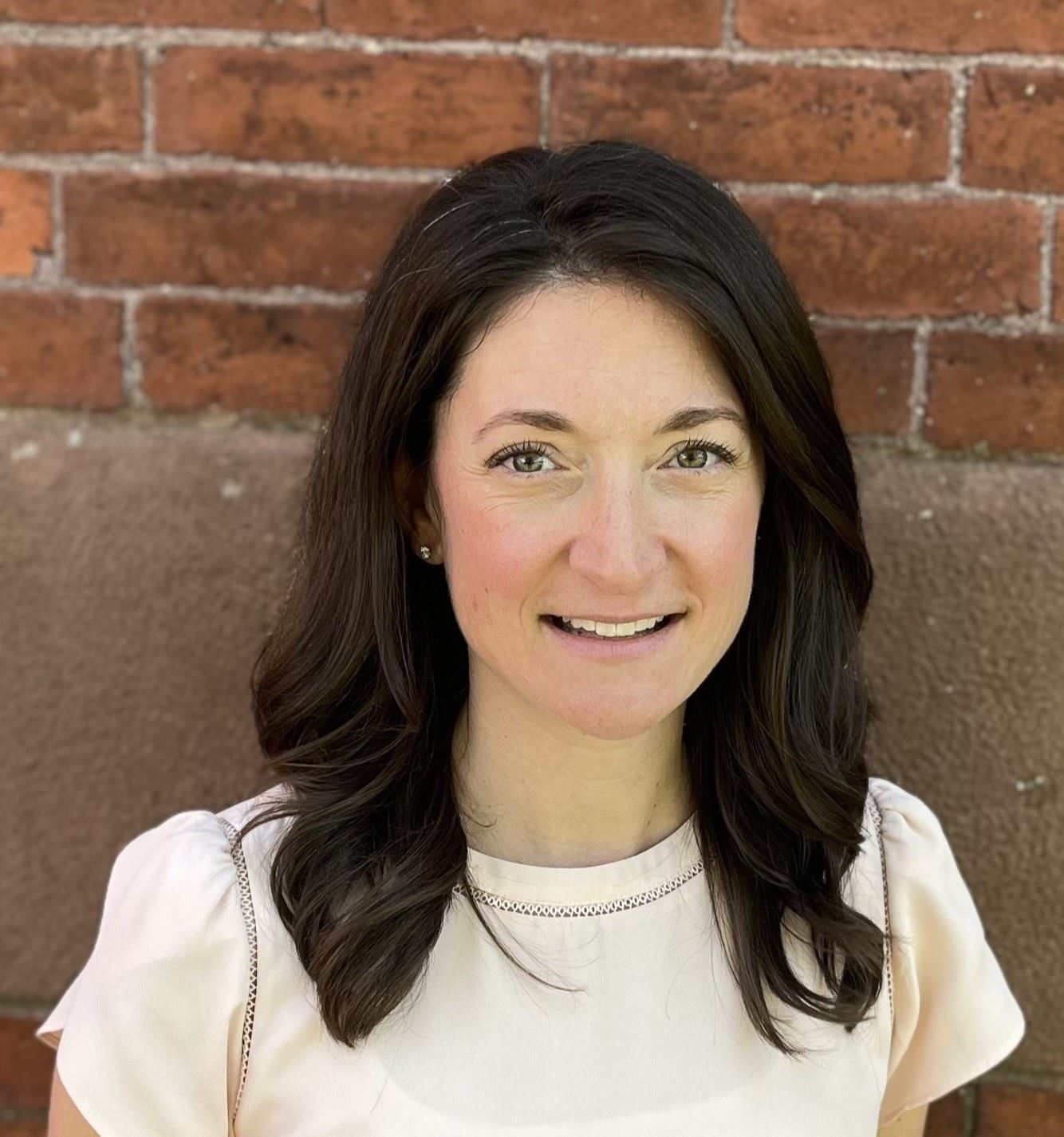 Woman with brown hair and white shirt smiling, in front of a brick background